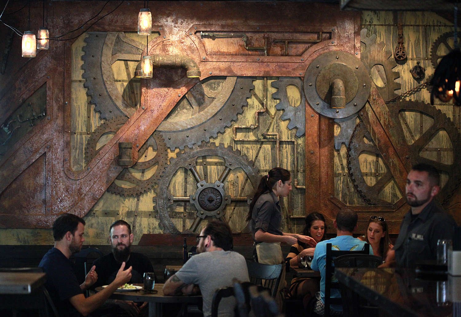Diners at tables in front of a wall decorated with industrial gears and pipes