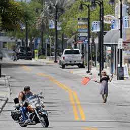A motorcyclist rides down a two-lane street lined with quaint shops and restaurants, looking over his shoulder at the only other visible person, a woman trying to attract customers by waving a red flag.